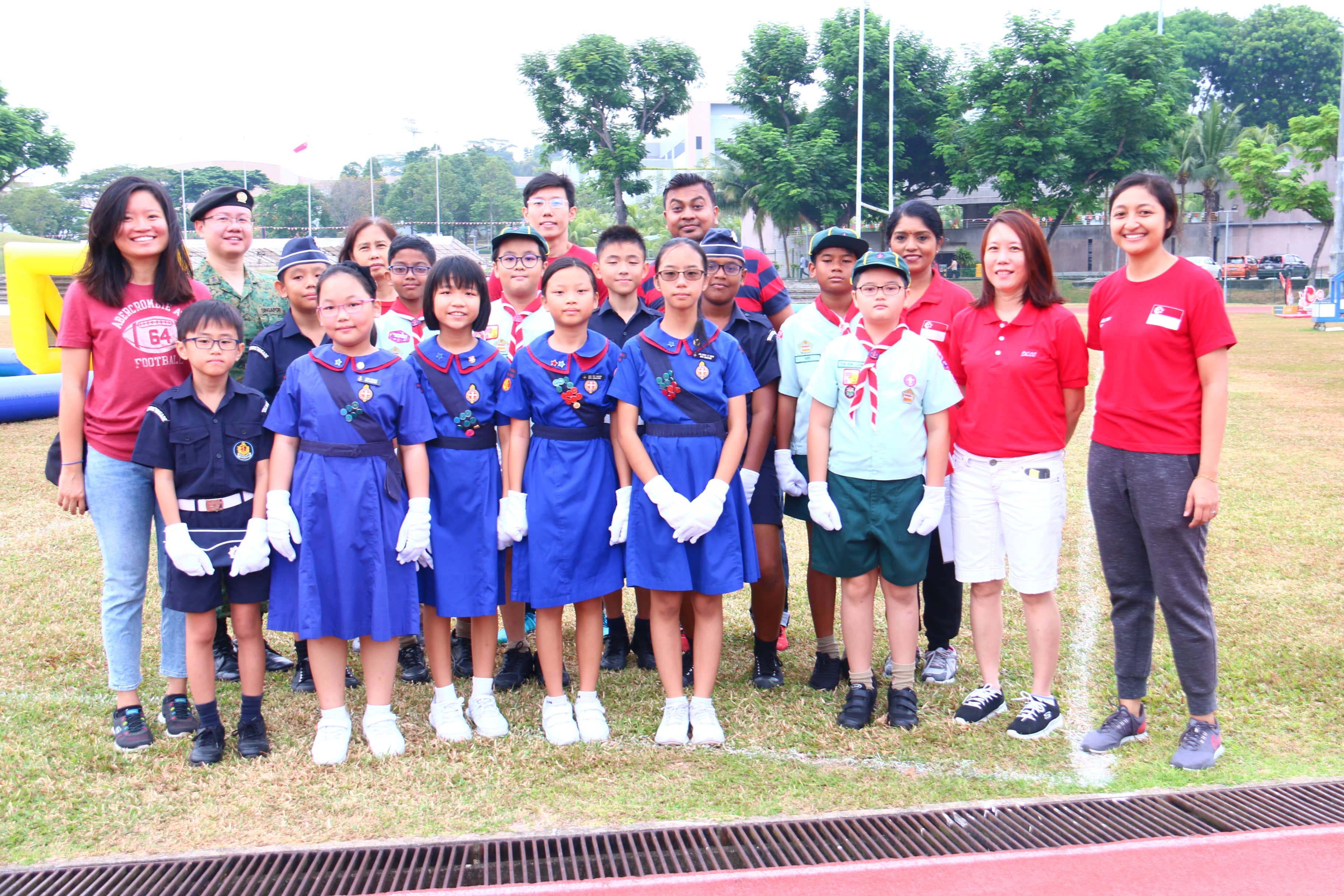 A group of children in scout and cadet uniforms pose with adults on a grassy field outdoors.